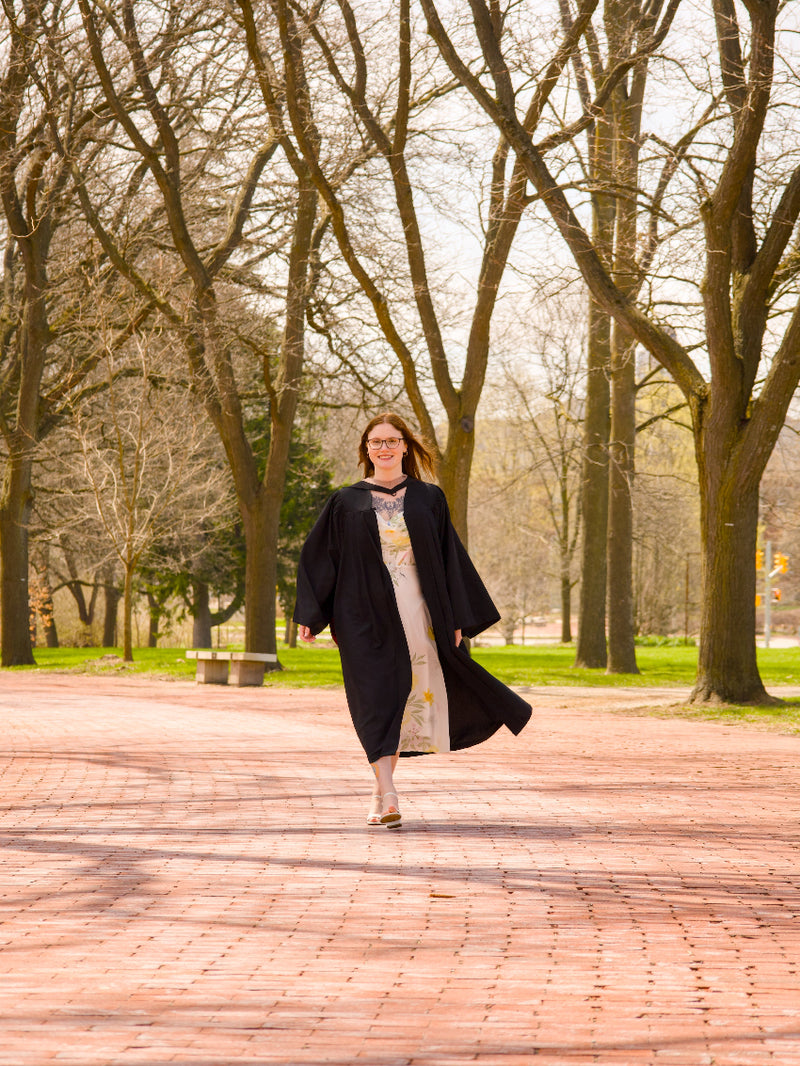 A professional photo of a Guelph grad on the University of Guelph campus in a black graduation gown, walking on a brick pathway of Alumni Walk. The photo is from Guelph Grad Photos.