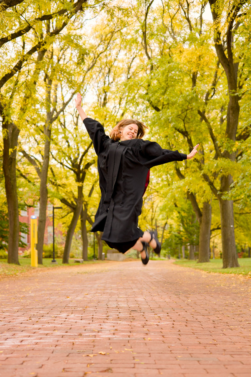 A professional photo of a Guelph grad in a black graduation gown jumping in a park with trees and a brick path. The photo is from Guelph Grad Photos. Edited