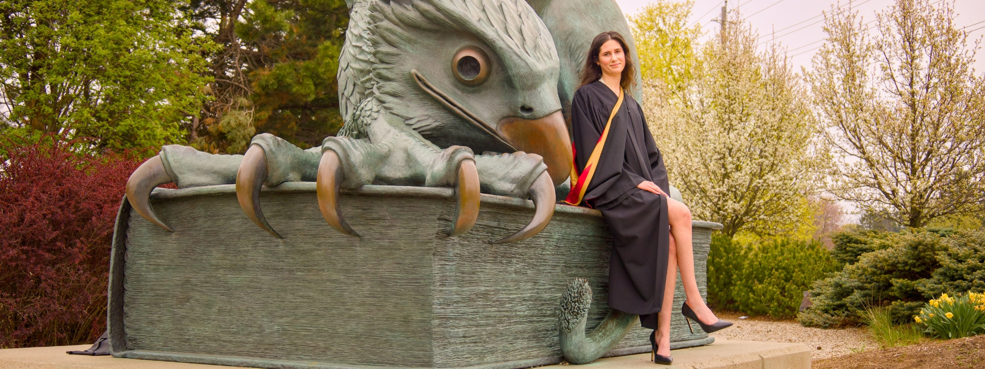 A woman in a graduation gown sitting on the Guelph Gryphon sculpture on Campus