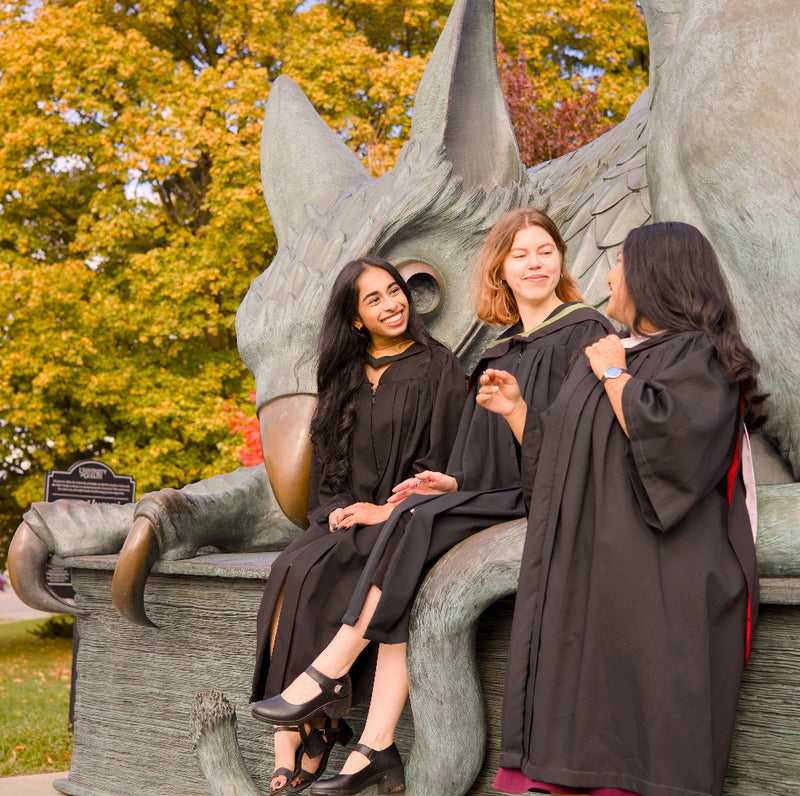 A professional photo of three Guelph grads on the University of Guelph campus. individuals in graduation gowns sitting on The Gryphon Statue on the University of Guelph Campus with autumn trees in the background.