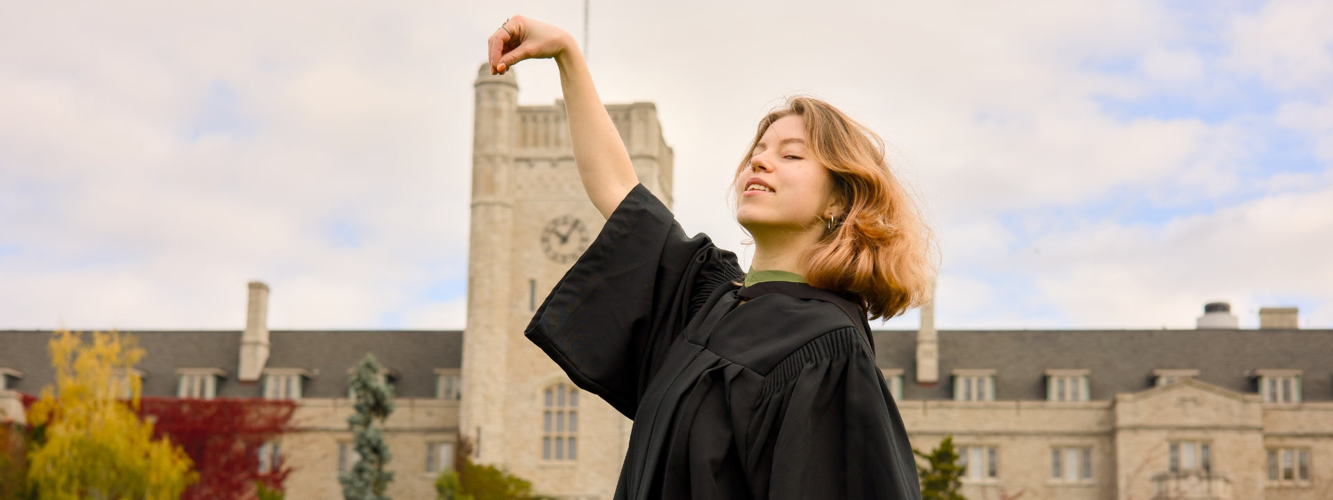 A Guelph Grad posing for photos in a graduation gown, standing in front of the Guelph University building with a clock tower.