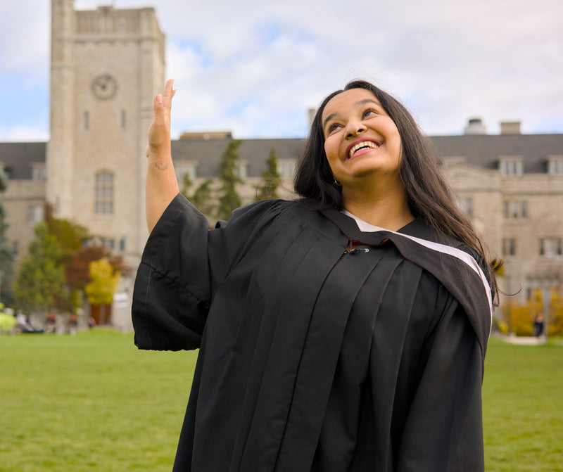 Graduate in a black cap and gown waving outdoors with a building in the background. The photo is from Guelph Grad Photos. The background is the University of Guelph Campus.
