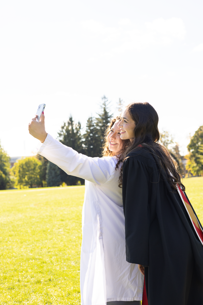 Two people taking a selfie outdoors in a park with trees and grass. The photo is from Guelph Grad Photos. The background is the University of Guelph Campus.
