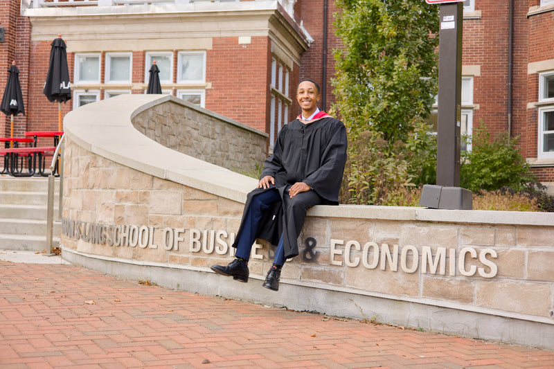 A professional photo of a Guelph grad on the University of Guelph campus in a cap and gown, sitting on a ledge with 'Economics' and 'School of Business' signs in the background. near the University of Guelph's LANG Building. The photo is from Guelph Grad Photos.