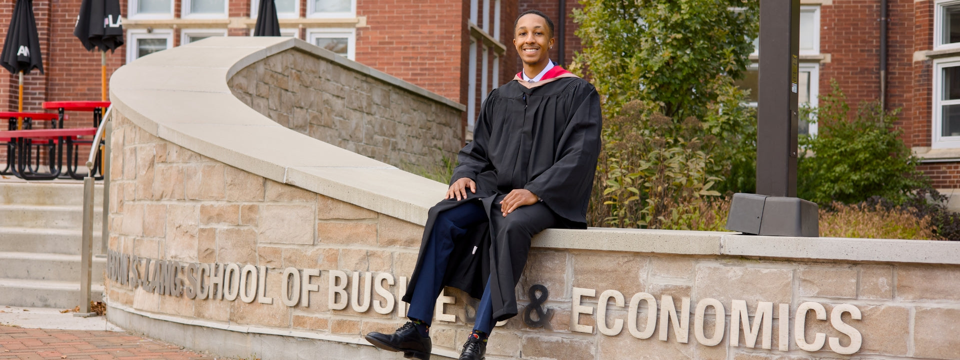 a Guelph grad in graduation gown sitting on a stone wall with 'LANG School of Business & Economics' inscription.