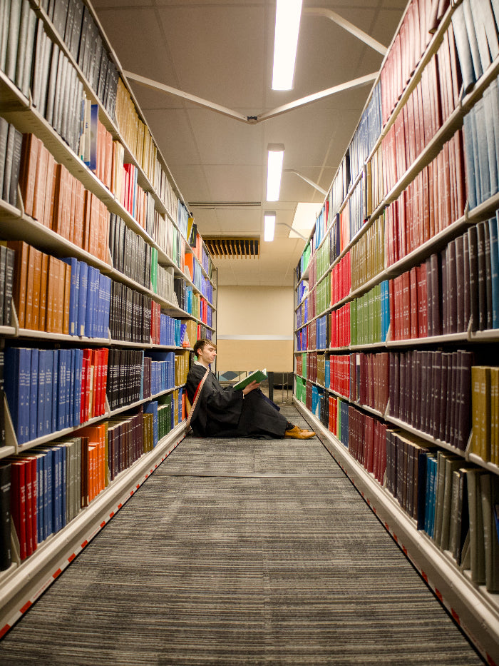 a Guelph Grad sitting on the floor between two rows of bookshelves in the McLaughlin Library. The photo is from Guelph Grad Photos.