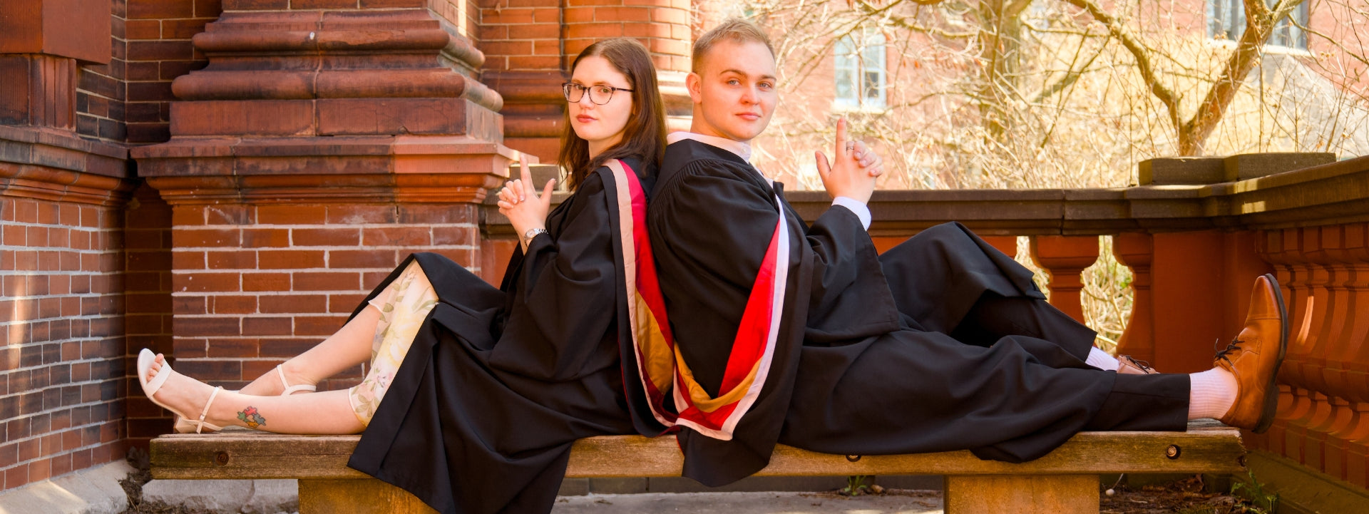  A professional photo of two young Guelph grads on the University of Guelph campus in black graduation gowns with red and yellow trim, sitting on a bench near the Macdonald Institute on the University of Guelph's campus with trees in the background. 
