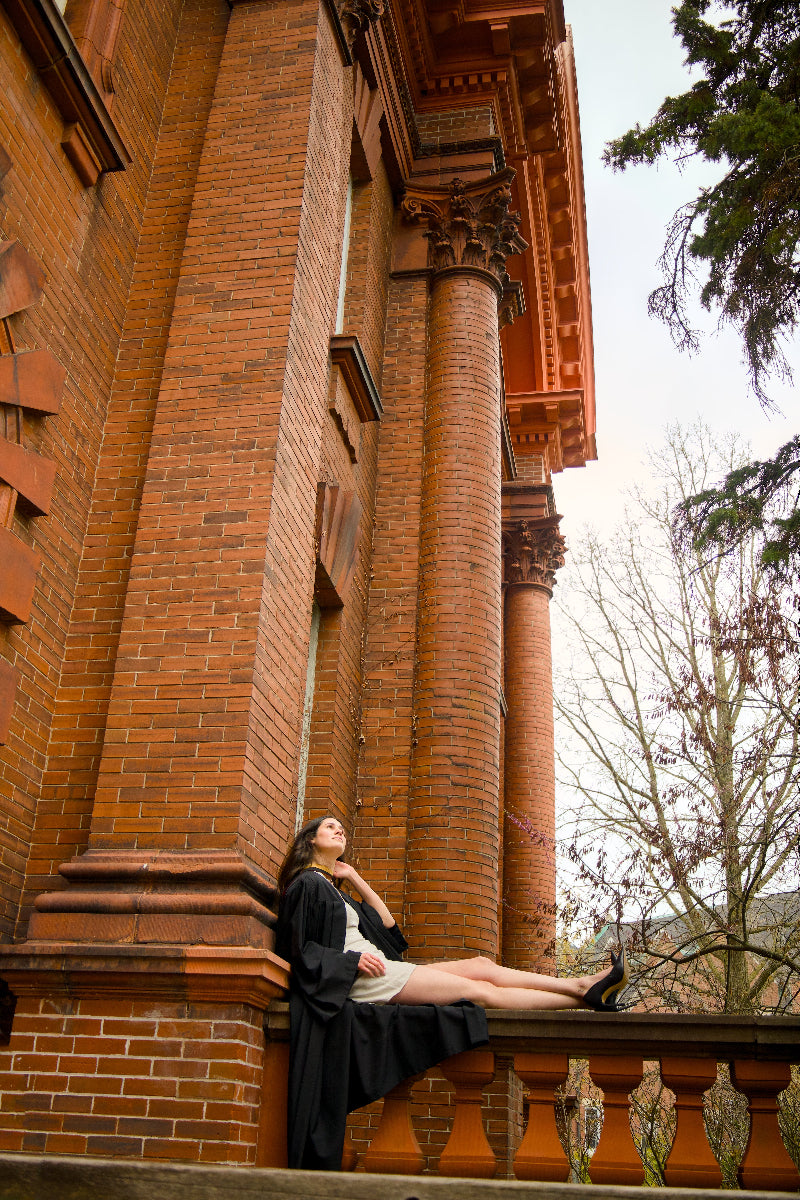 Woman in a black dress sitting on a stone ledge against a brick building with columns. The photo is from Guelph Grad Photos. The background is the University of Guelph Campus. 