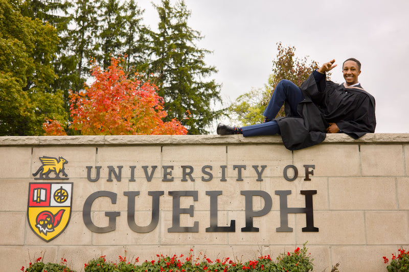 Person in graduation gown sitting on the University of Guelph sign with trees and flowers in the background. The photo is from Guelph Grad Photos. The background is the University of Guelph Campus.