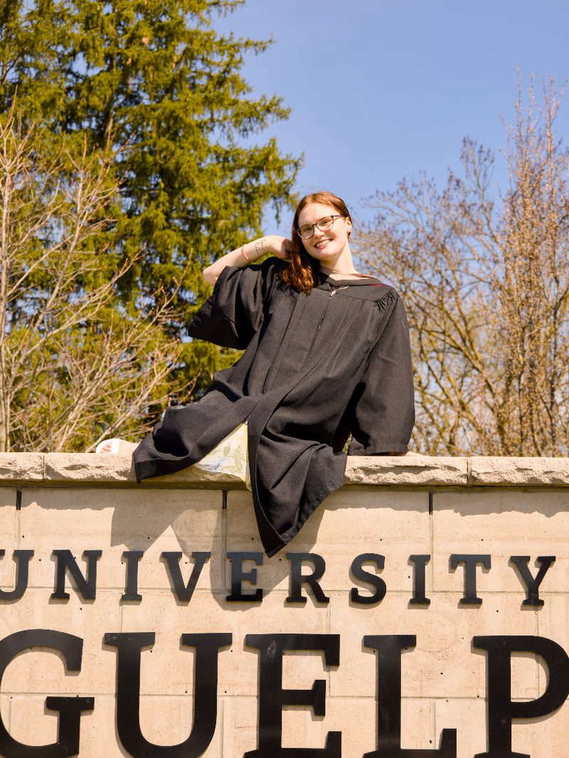 Person in graduation gown sitting on a stone wall with 'University of Guelph' sign in the background, The photo is from Guelph Grad Photos. The background is the University of Guelph Campus.