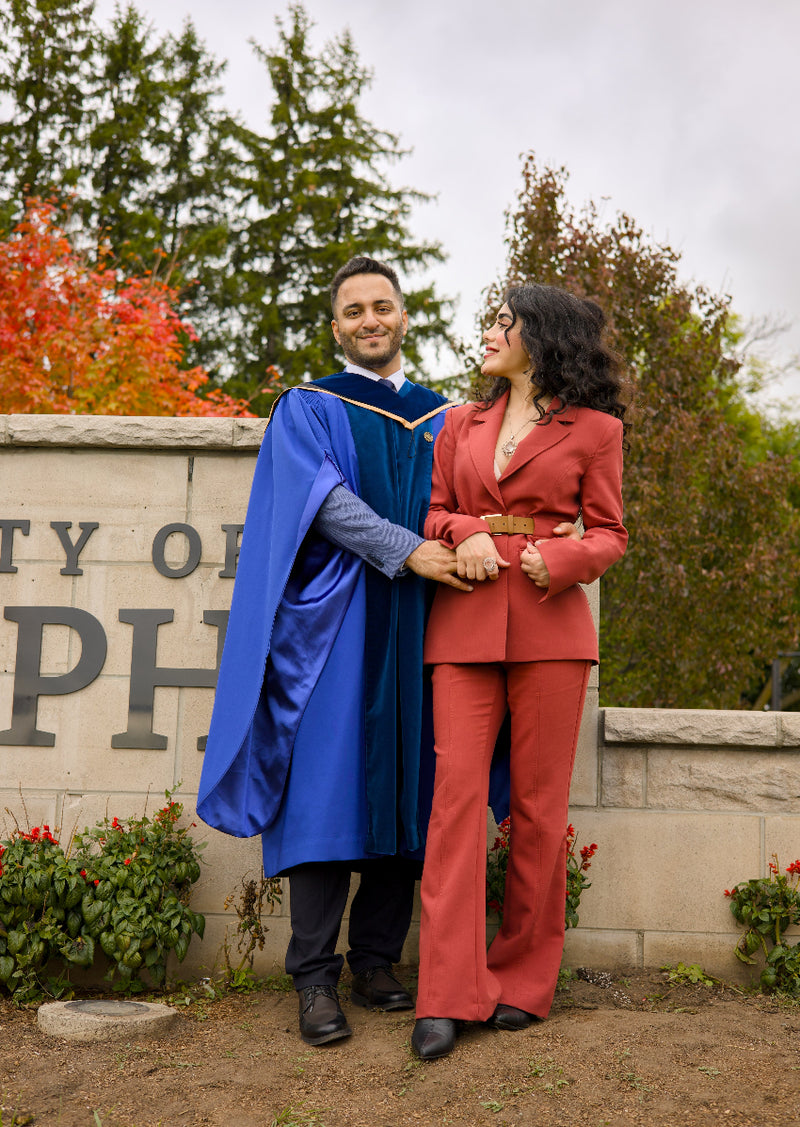 Graduates in blue and red caps and gowns standing outdoors with a city sign in the background. The photo is from Guelph Grad Photos. The background is the University of Guelph Campus.
