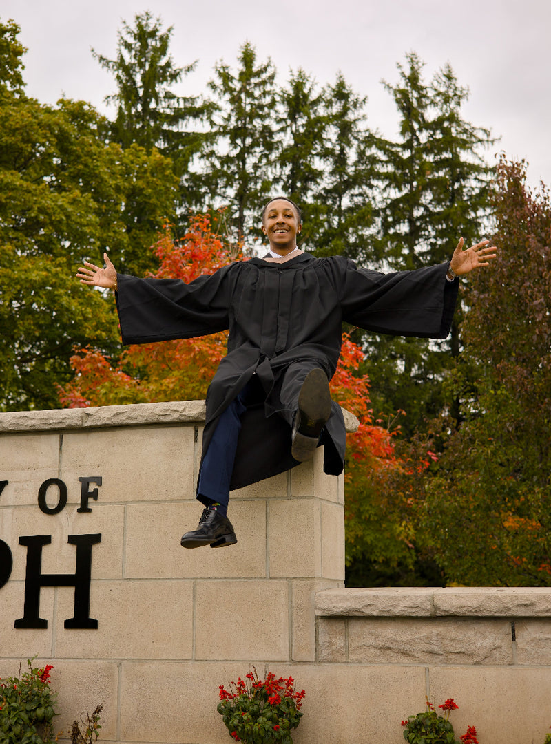 Graduate in cap and gown sitting on a stone wall with trees and 'University of ...' sign in the background. The photo is from Guelph Grad Photos. The background is the University of Guelph Campus.