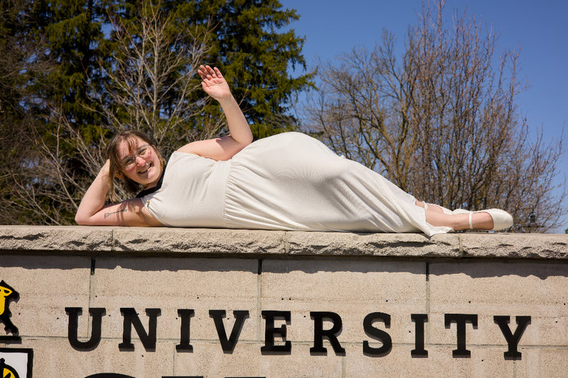 Person in a white dress lying on a stone sign with 'UNIVERSITY' at a university campus. The photo is from Guelph Grad Photos. The background is the University of Guelph Campus.
