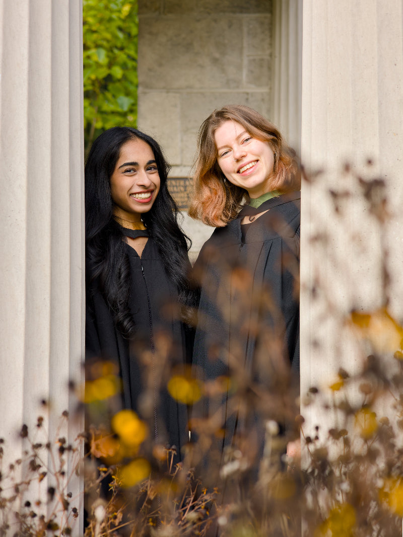 Two women standing behind a stone wall with autumn leaves and flowers in the foreground. The photo is from Guelph Grad Photos. The background is the University of Guelph Campus.
