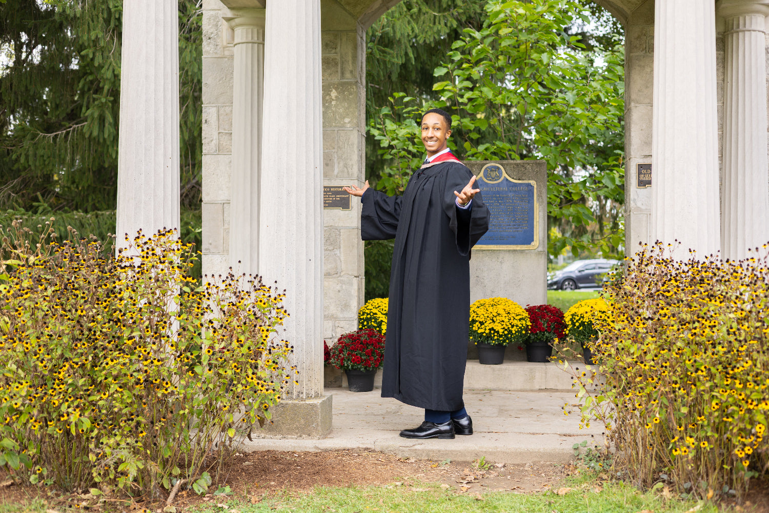 Graduate in a cap and gown standing in front of a classical building with columns and flowers. The photo is from Guelph Grad Photos. The background is the University of Guelph Campus.
