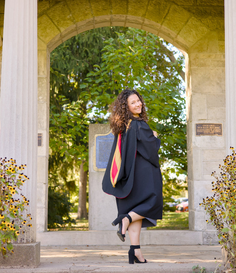 Woman in a graduation gown standing under an archway with greenery in the background. The photo is from Guelph Grad Photos. The background is the University of Guelph Campus, The Portico.