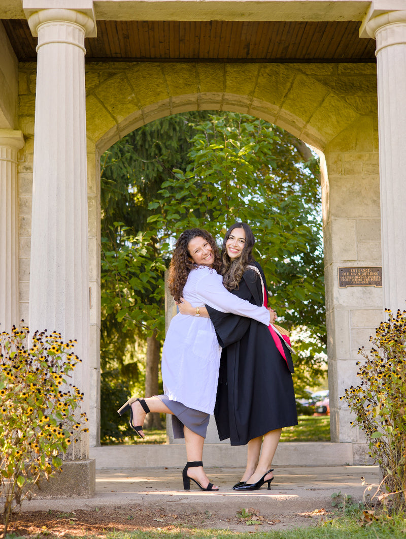 Two women hugging outdoors under a stone archway with greenery in the background. The photo is from Guelph Grad Photos. The background is the University of Guelph Campus.