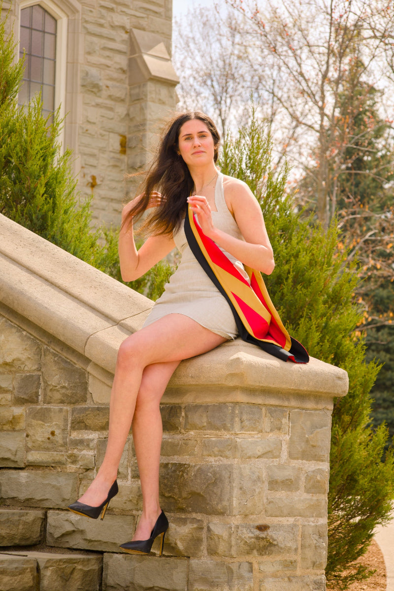 A Guelph grad posing for her graduation photo, sitting on stone steps, on the University of Guelph's campus, holding a sash with the war memorial building and trees in the background.