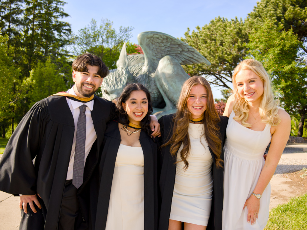 Four graduates in regalia laughing together in front of the Gryphon statue at the University of Guelph