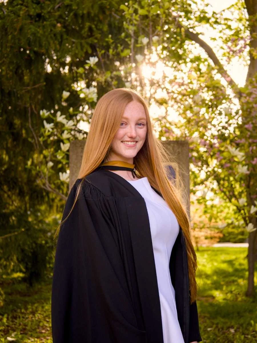A professional photo of a Guelph grad on the University of Guelph campus, graduating in a gown, standing outdoors with trees and sunlight in the background. The photo is from Guelph Graduation Photos. The background is the University of Guelph Campus.