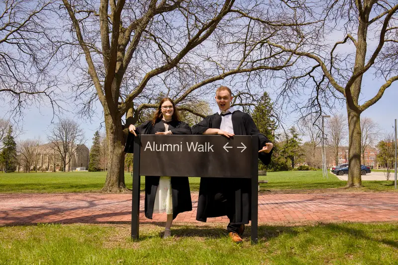 Two Guelph Grads in graduation gowns holding an 'Alumni Walk' sign outdoors.