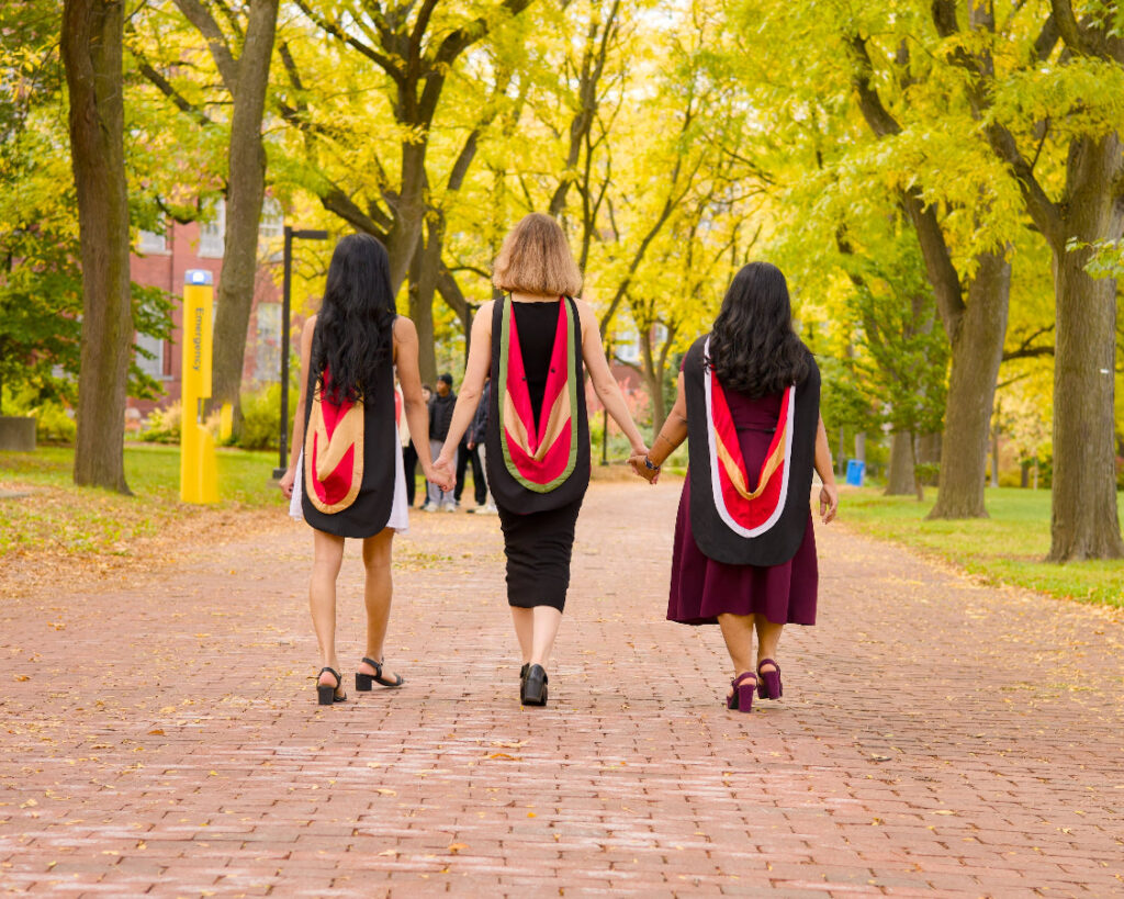 Three graduates walking hand in hand down the brick path of Alumni Walk under yellow autumn trees, stoles across their backs