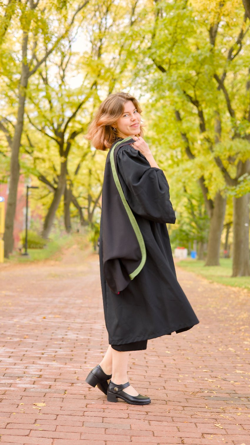 A professional photo of a young Guelph grad on the University of Guelph campus in a black graduation gown with green trim, standing on a brick path on the Alumni Walk pathway on the University of Guelph's campus with trees in the background.