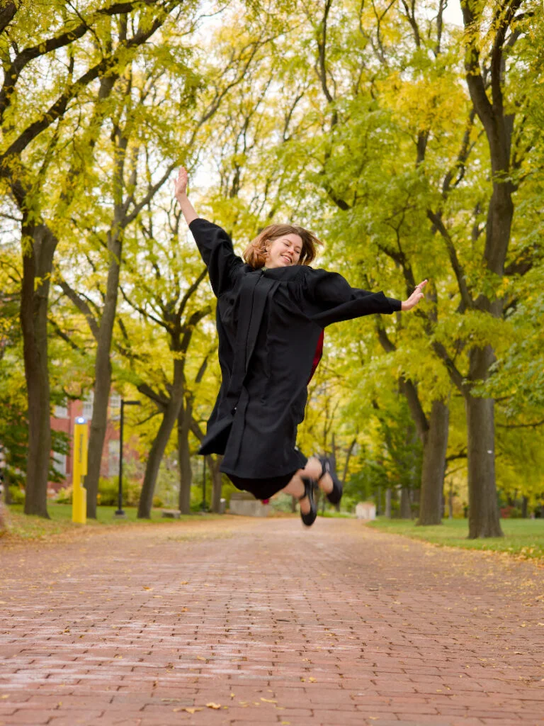 A professional photo of a young Guelph grad on the University of Guelph campus wearing graduation gown and hood. She is jumping joyfully on the Alumni Walk pathway on the University of Guelph's campus with trees in the background. The photo is from Guelph Grad Photos.