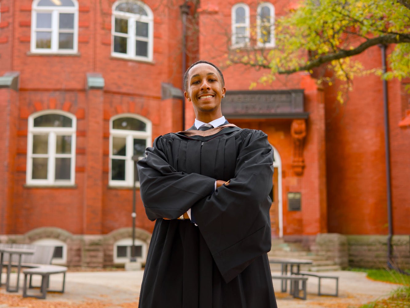 Graduate in black cap and gown standing in front of a red brick building with trees in the background.