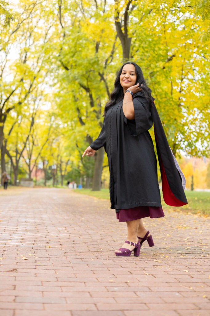 A young woman in graduation gown, on Alumni Walk on the University of Guelph Campus, with green trees in the background with trees in the background. The photo is from Guelph Grad Photos.