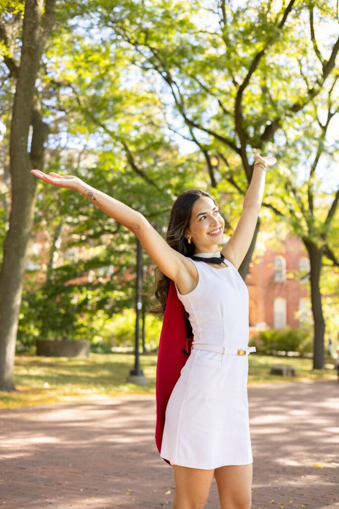 A young woman in a white dress, on Alumni Walk on the University of Guelph Campus, with green trees in the background with trees in the background. The photo is from Guelph Grad Photos.