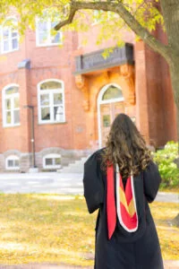 A young womanin graduation gown with a red and yellow stole standing in front of a brick building. on Alumni Walk, a tree-lined path on the University of Guelph Campus, with a brick building. in the background. The photo is from Guelph Grad Photos.