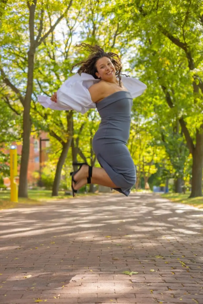 Woman in a gray dress jumping in a park with trees and a path in the background