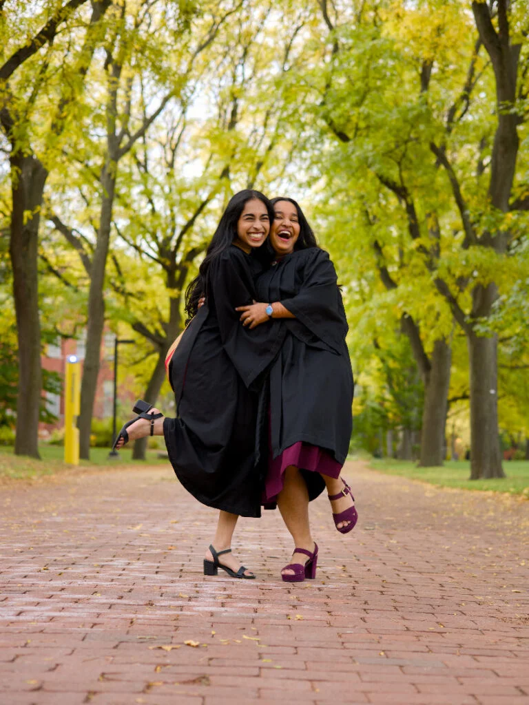 Two women in graduation gowns posing together on a brick path with trees in the background.