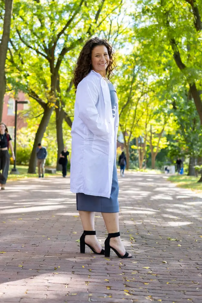 A young woman in a white lab coat standing on a brick path with trees and people in the background, on Alumni Walk Tree-Lined Path on the University of Guelph Campus, with autumn trees in the background. The photo is from Guelph Grad Photos.