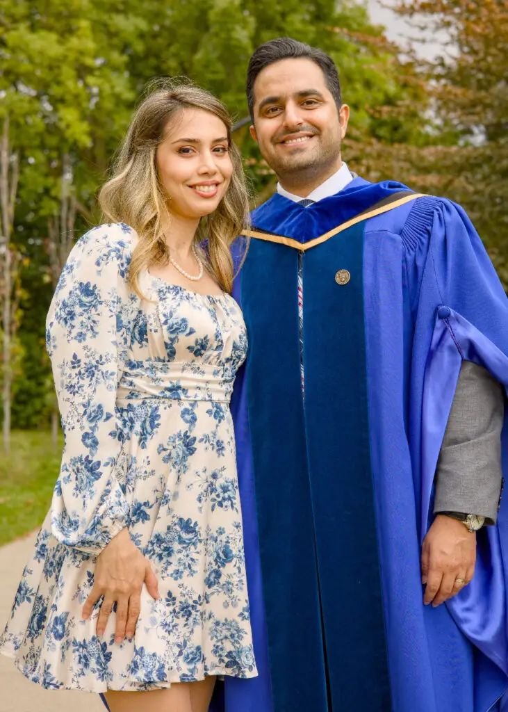 A professional photo of a Guelph PHD grad on the University of Guelph campus in a blue gown with a floral dress, and a person outdoors. The photo is from Guelph Grad Photos. The background is The Portico on the University of Guelph Campus.