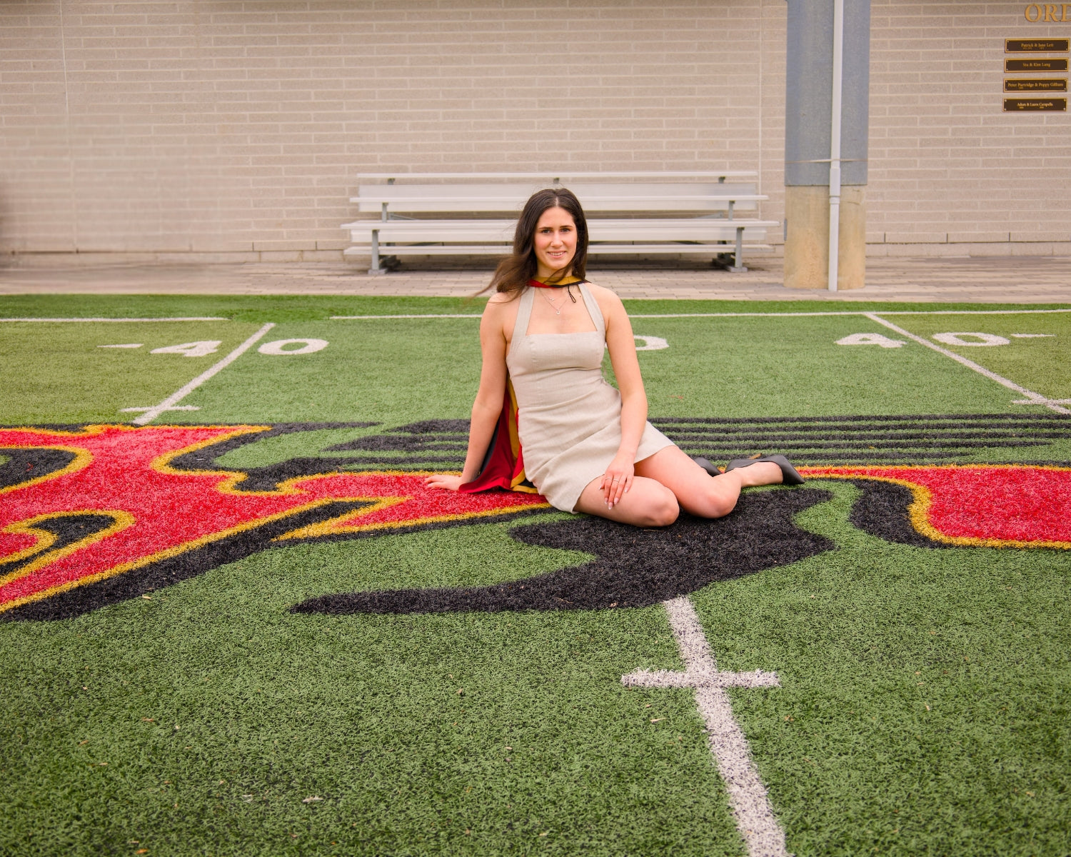 Woman in a white dress sitting on a sports field with a bench in the background. The photo is from Guelph Grad Photos. The background is the University of Guelph Campus.