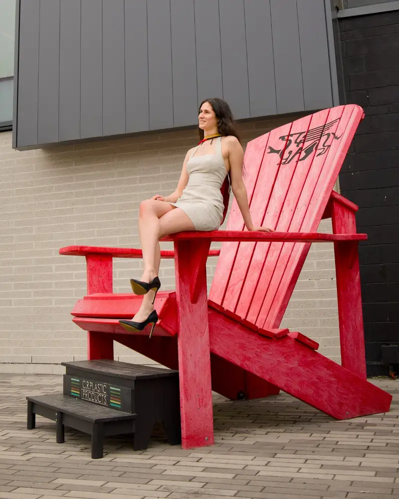 a Guelph grad on the University of Guelph campus, sitting on a large red chair with a black design, outdoors. The photo is from Guelph Grad Photos. The background is the Gryphon Alumni Stadium on the University of Guelph Campus.
