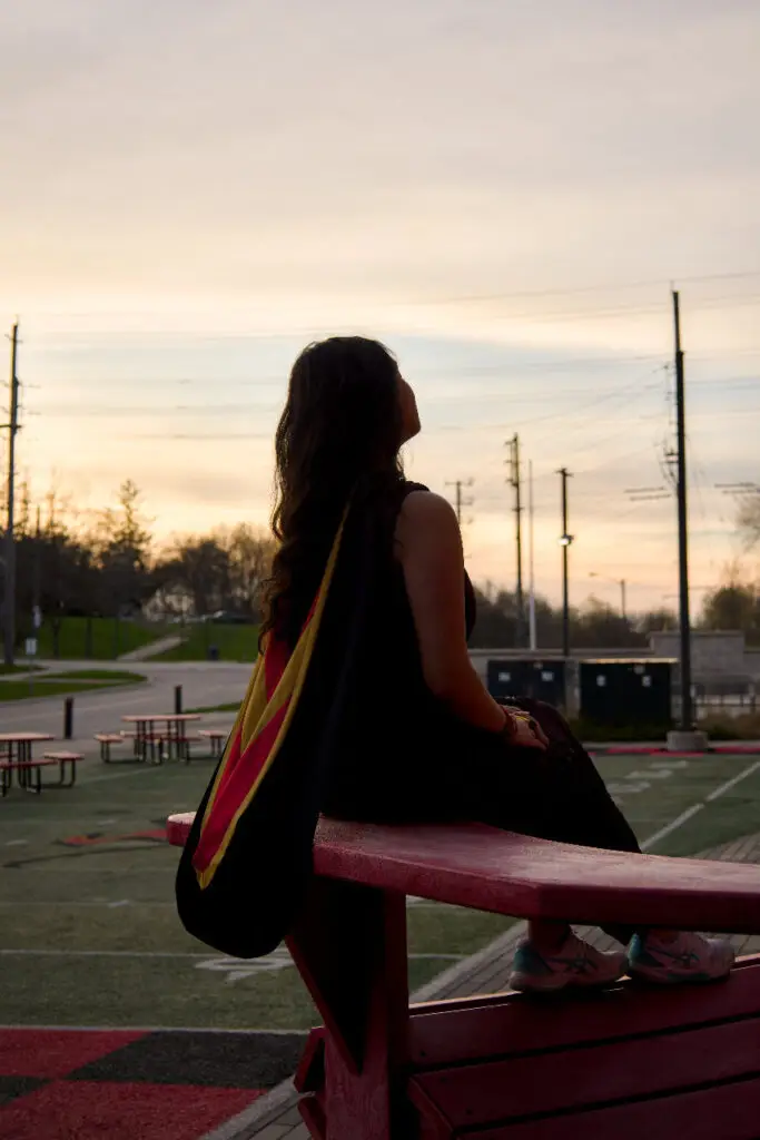 A person sitting on a bench with a flag, looking at a sunset over an empty parking lot. The photo is from Guelph Grad Photos. The background is the University of Guelph Campus.