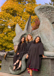 Three young women in graduation gowns sitting on The Gryphon Statue on the University of Guelph Campus, with autumn trees in the background. The photo is from Guelph Grad Photos.