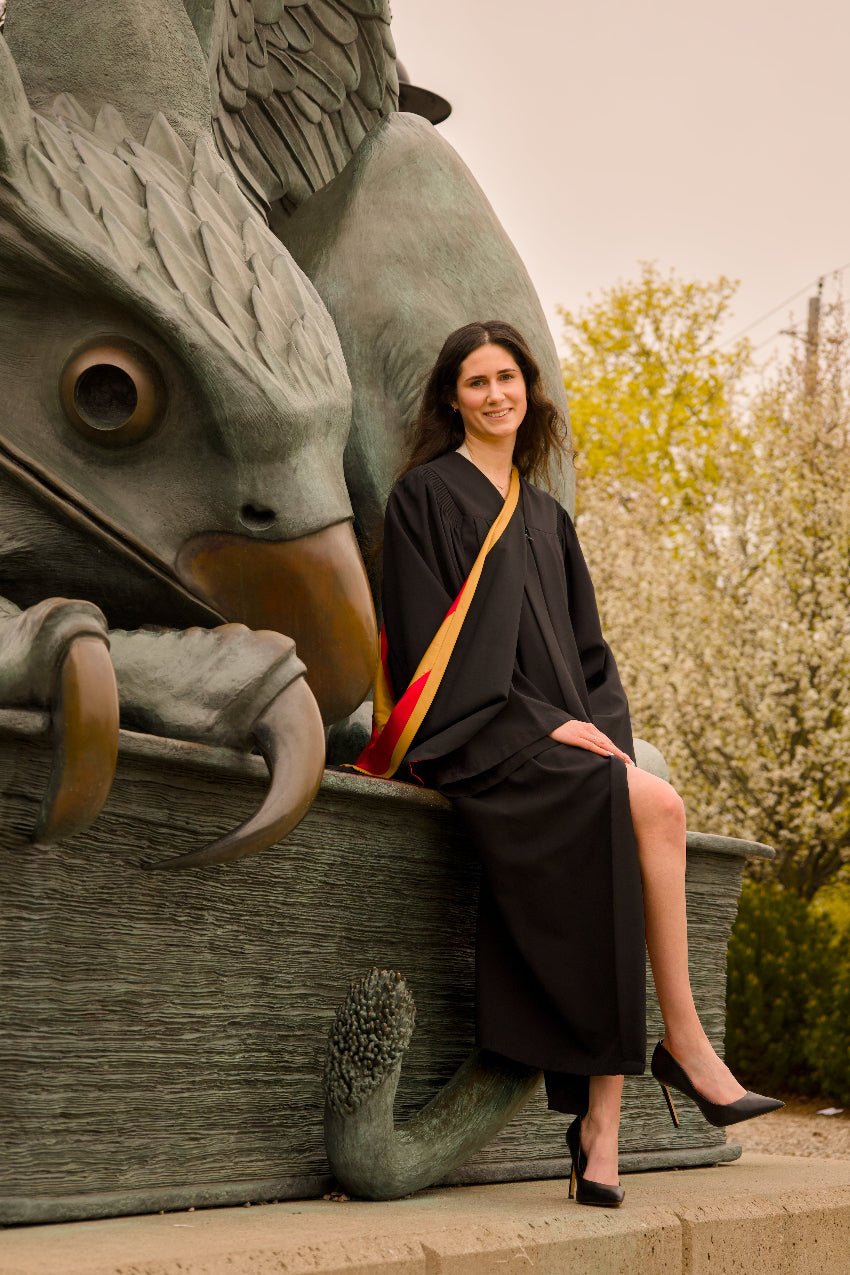 Woman in graduation gown sitting on a stone lion sculpture with trees in the background. The photo is from Guelph Grad Photos. The background is the University of Guelph Campus.