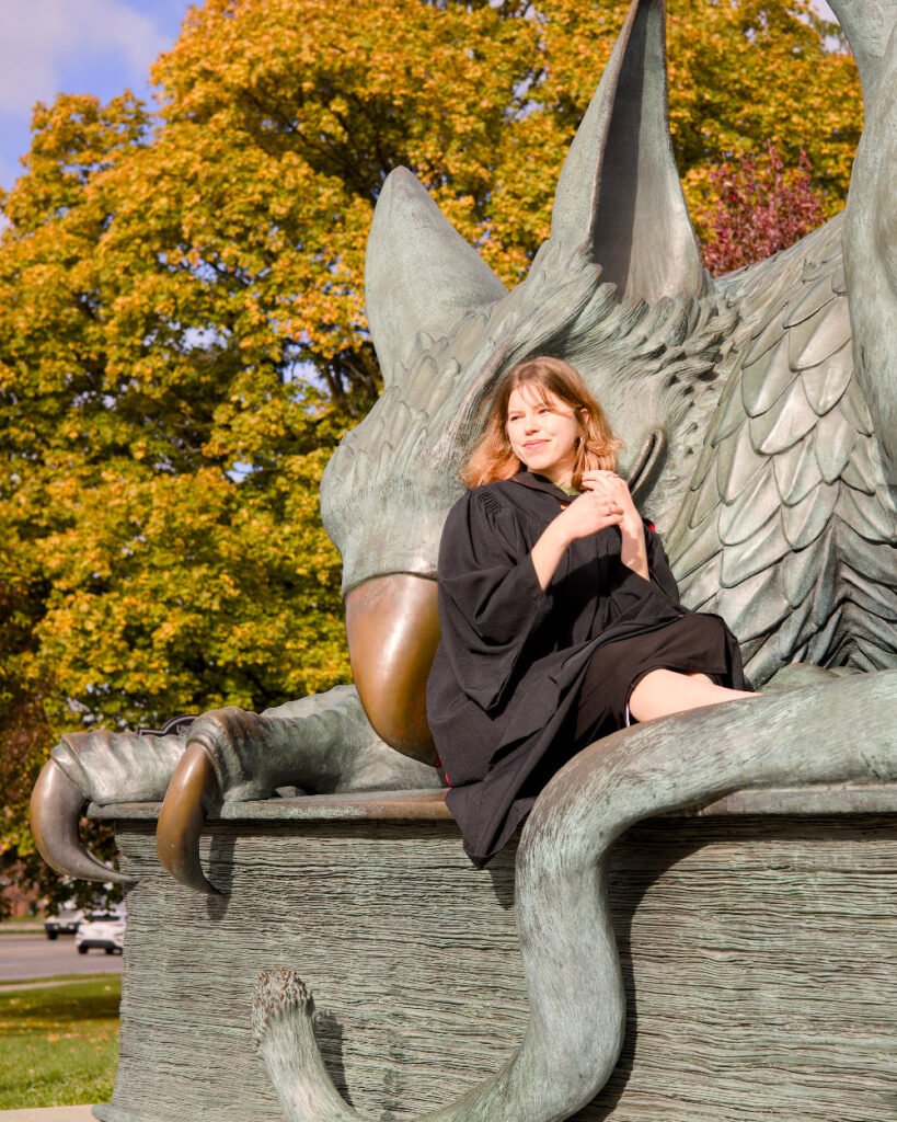 A professional photo of a young woman in a graduation gown. sitting on the Gryphon Statue on the University of Guelph Campus. The photo is from Guelph Grad Photos.