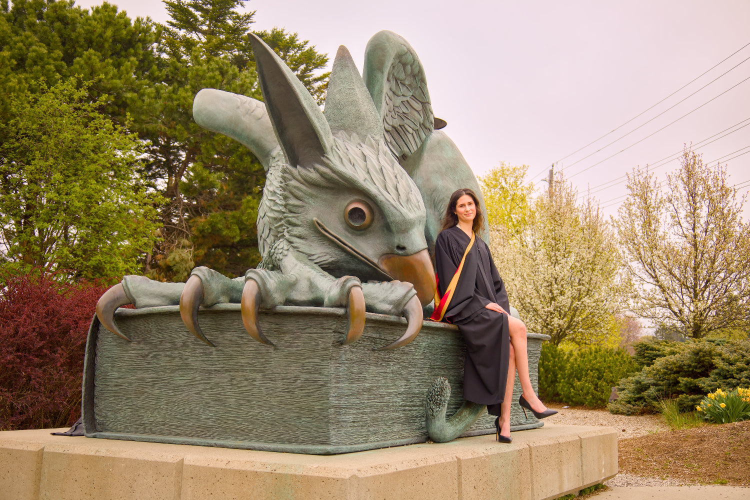 Woman in graduation gown sitting on the gryphon statue outdoors. The photo is from Guelph Grad Photos. The background is the University of Guelph Campus.