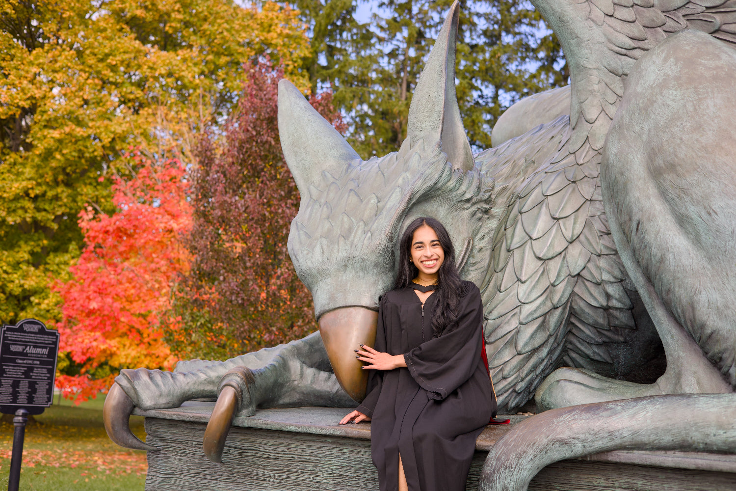 a Guelph Grad posing with the Guelph Grayphon statue in an outdoor setting with trees displaying autumn colours. The photo is from Guelph Grad Photos. The background is the University of Guelph Campus.