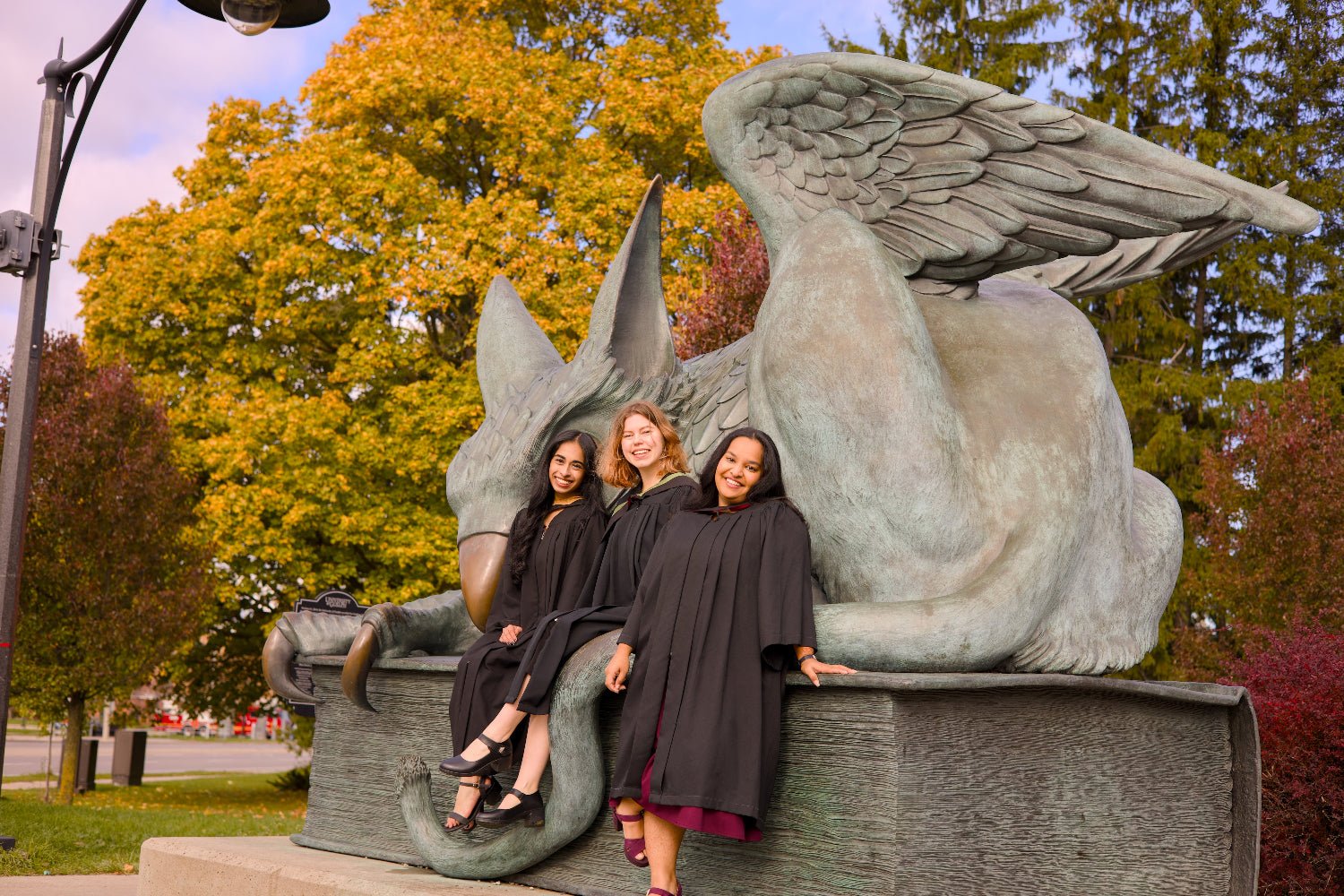 Three women in graduation gowns posing on the gryphon statue on campus. The photo is from Guelph Grad Photos. The background is the University of Guelph Campus.