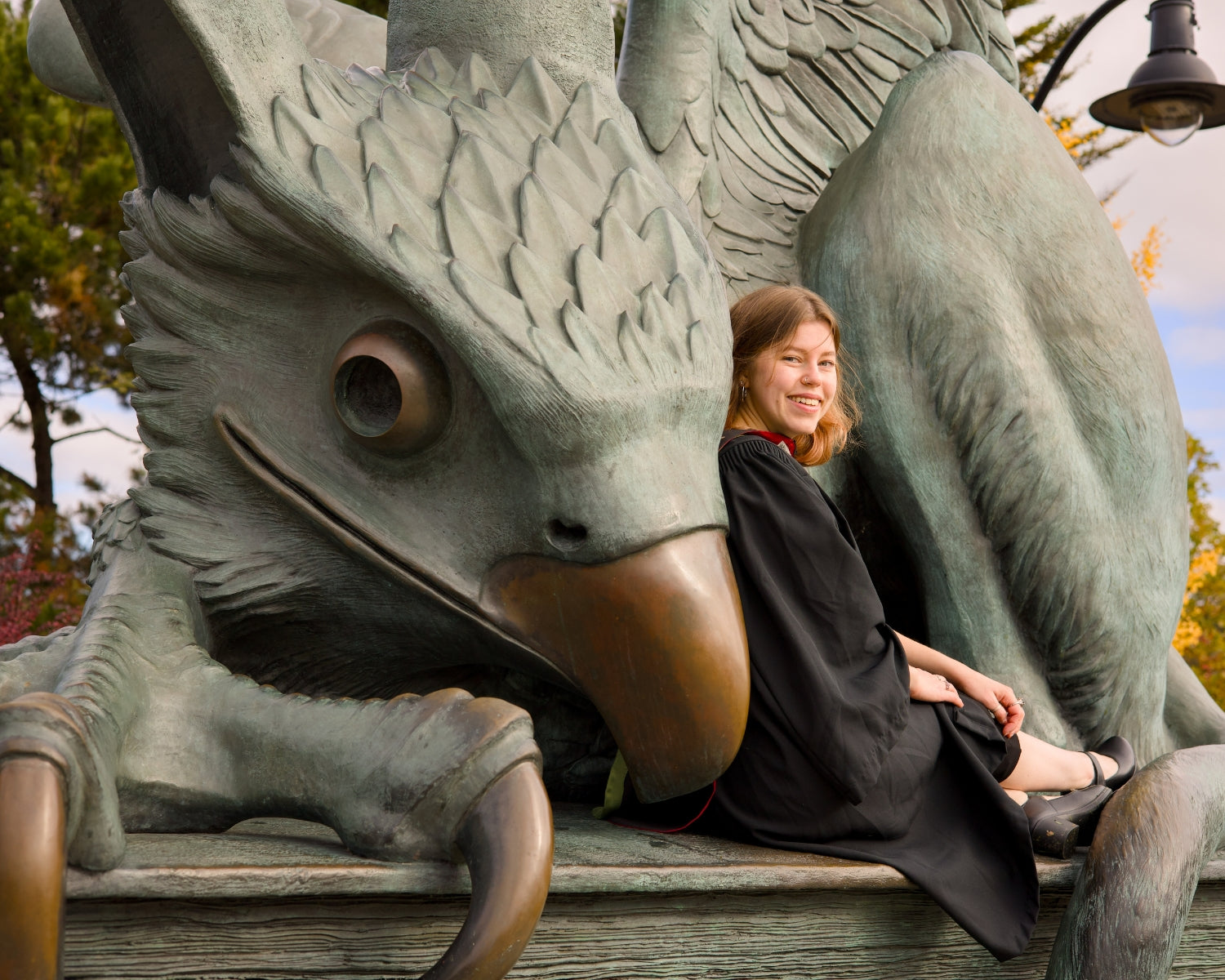 A professional photo of a Guelph grad on the University of Guelph campus, she is sitting on the Guelph Gryphon statue with trees and sky in the background. The photo is from Guelph Grad Photos. The background is the Gryphon Statue on the University of Guelph Campus.
