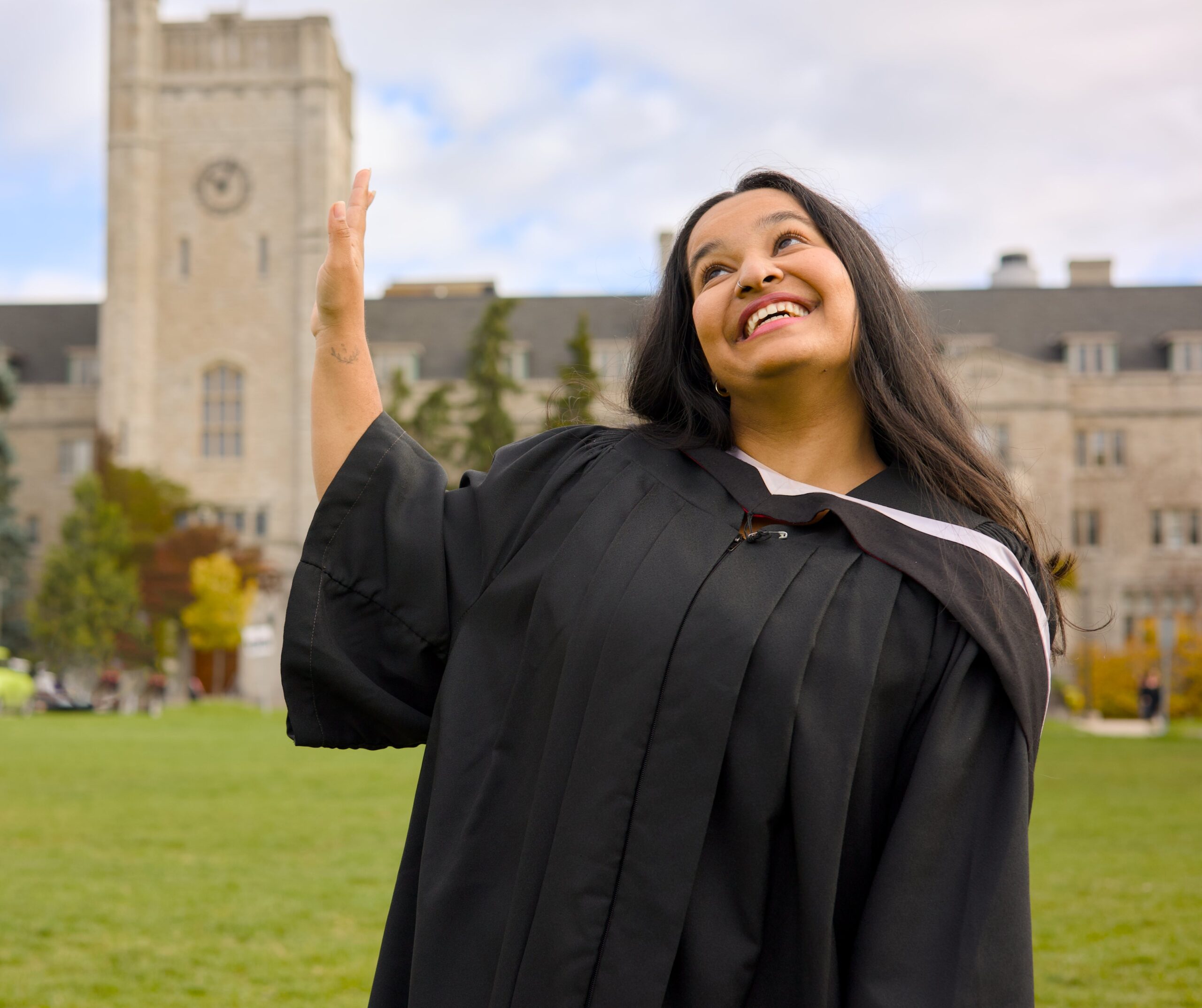 Graduate in a black cap and gown waving outdoors with a building in the background. The photo is from Guelph Grad Photos. The background is the University of Guelph Campus.