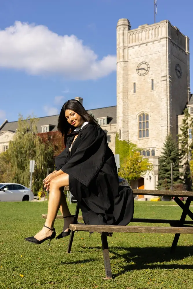 A University of Guelph graduate in cap and gown sitting on a bench in front of the Johnston Hall clock tower