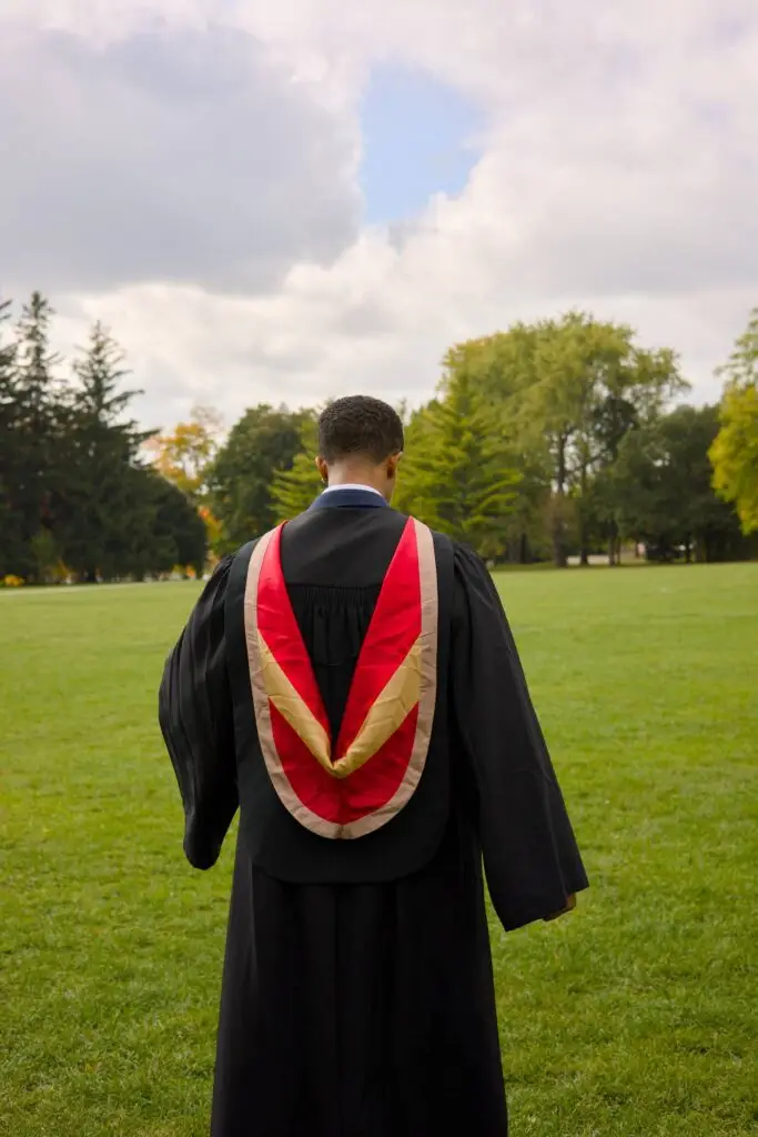 Person in a graduation gown with a red and gold stole standing on a grassy field with trees in the background.