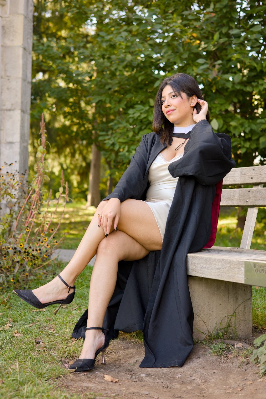 Person in graduation gown sitting on a bench outdoors with trees in the background. The photo is from Guelph Grad Photos. The background is the University of Guelph Campus.
